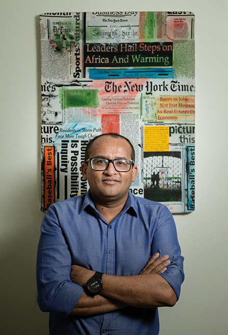 Fotografia mostra, em primeiro plano, o professor Francivaldo Alves Nunes. Ele usa camisa azul, tem cabelos pretos e curtos, está de óculos e sorri em nossa direção. Atrás dele, um quadro colorido está pendurado na parede.